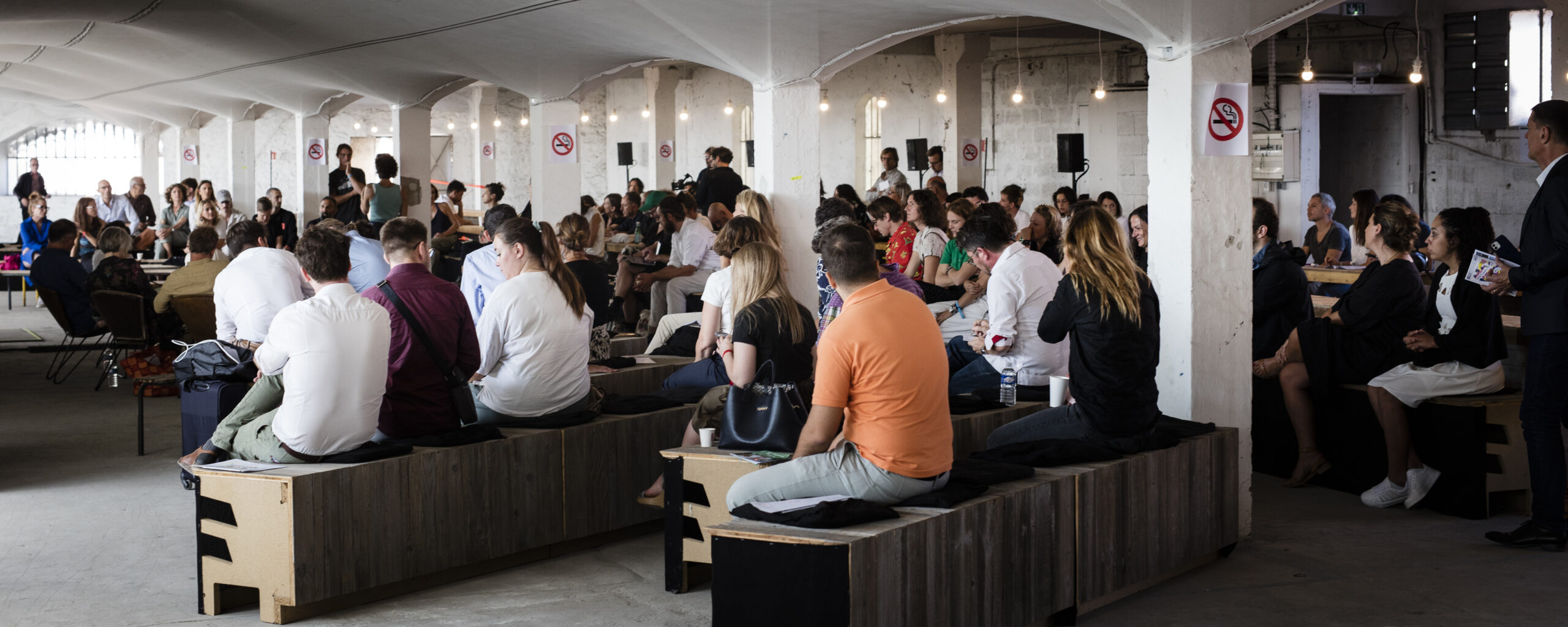 Public assis sur des gradins en bois sous les voûtes du site industriel, lors d'une conférence du Festival de la Ville Sauvage à Marseille